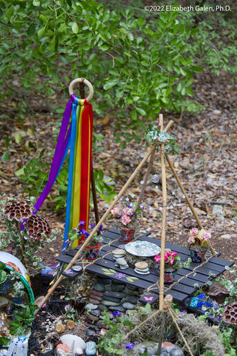 A miniature picnic table with skewer bowers and stone plates surrounded by tiny flowers and a rainbow ribbon windsock